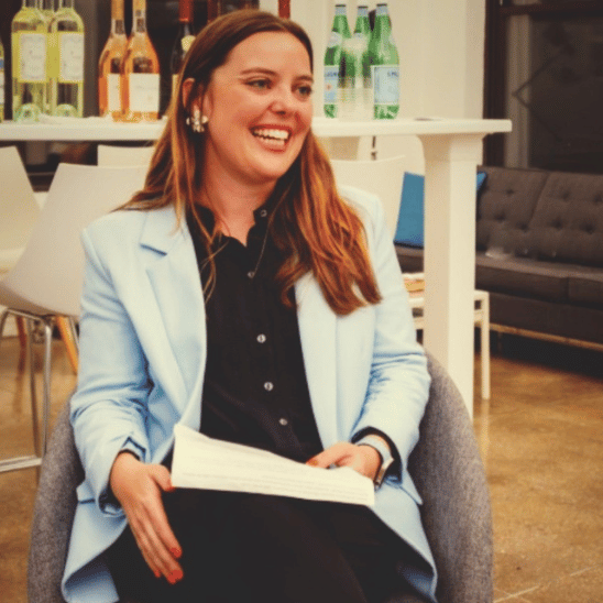 Smiling woman in a light blue blazer seated with a paper in hand, in a casual event space with drinks on a shelf behind her.
