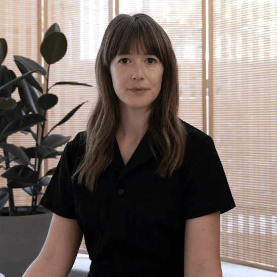 Portrait of a woman with shoulder-length brown hair wearing a black shirt, seated indoors with blinds and a plant in the background.