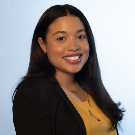 Portrait of a smiling woman with long dark hair, wearing a black blazer and yellow blouse against a light blue background, suitable as a professional headshot.