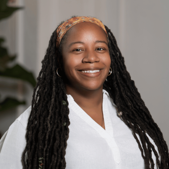 Smiling woman with long dreadlocks, wearing a peach headband and white blouse, against a neutral background.