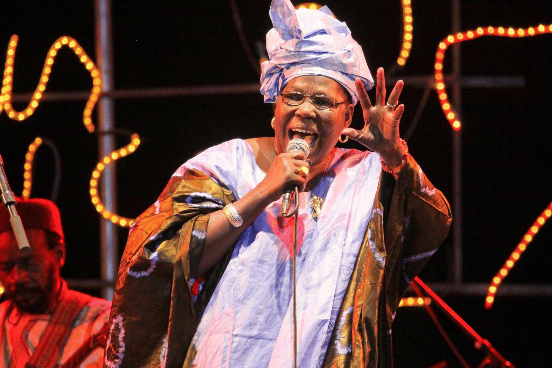 A performer in a flowing light-colored robe and matching headwrap sings into a handheld microphone on a stage. One hand is raised in an expressive gesture. Warm orange stage lights form curved patterns in the dark background, while another musician is partially visible to the side.
